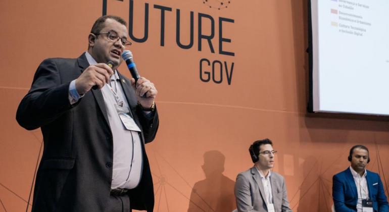 Homem de terno palestra, de pé; ao fundo, na parede, os dizeres: "Future gov". 