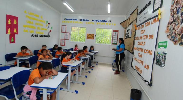 Oito alunos, sentados em suas carteiras, assistem a aula de professora. 