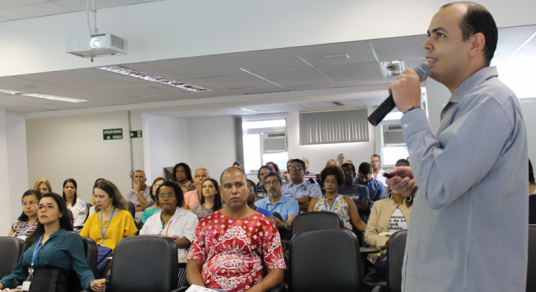 Fisioterapia preventiva com foco na coluna vertebral foi o tema de palestra oferecida pela Prefeitura na quarta-feira, dia 27/3. Foto: Divulgação PBH Homem, de pé, com microfone na mão, palestra para cerca de vinte pessoas, sentadas, ao fundo.