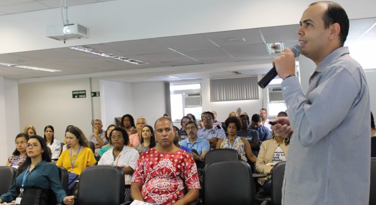 Homem, de pé, com microfone na mão, palestra para cerca de vinte pessoas, sentadas, ao fundo. 