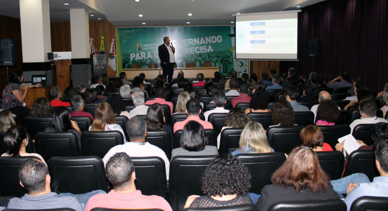 Homem, de pé, palestra para mais de cinquenta pessoas sentadas no salão JK, da Prefeitura. 