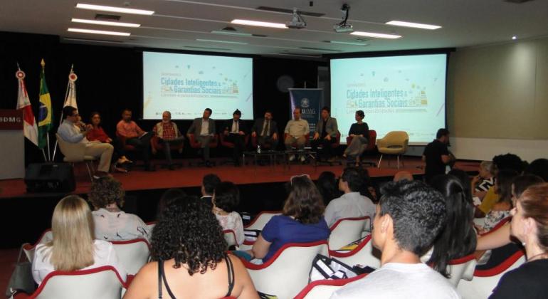 Mais de onze pessoas assistem a palestra com onse pessoas no palco e telões, em uma sala. 