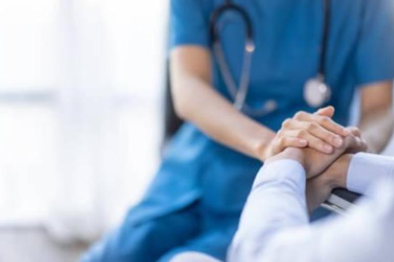 A imagem apresenta um médico que só aparece até a altura dos ombros. Está com roupa azul e estetoscópio sobre os ombros. Ele coloca as mãos sobre as mãos de outra pessoa que está de branco