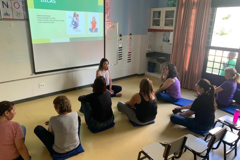 Pessoas sentadas no chão de uma sala com uma professora sentada à frente 