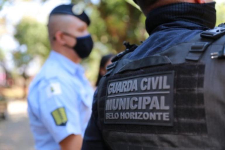 Dois guardas municipais um está de costas em primeiro plano mostrando somente o colete com a frase Guarda Civil Municipal de Belo Horizonte. Outro guarda está em segundo plano usando máscara 