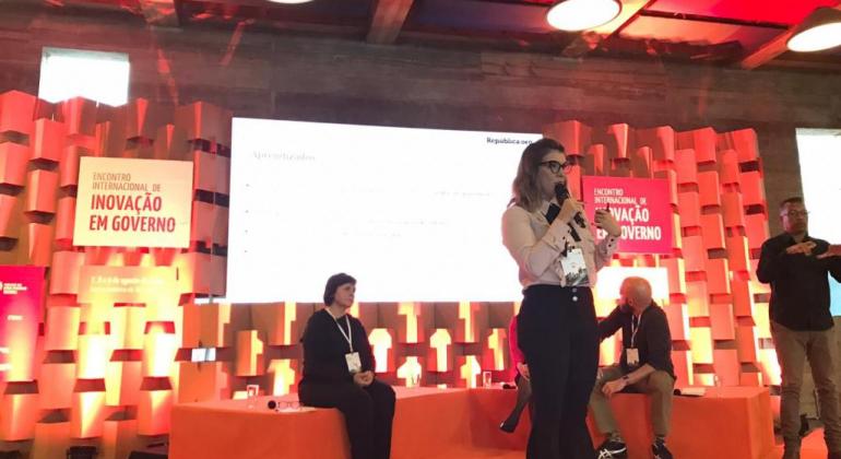 A iniciativa foi apresentada no painel “Táticas e estratégias de engajamento de servidores: relatos de experiências de gestão de pessoas”. Mulher palestra em evento.