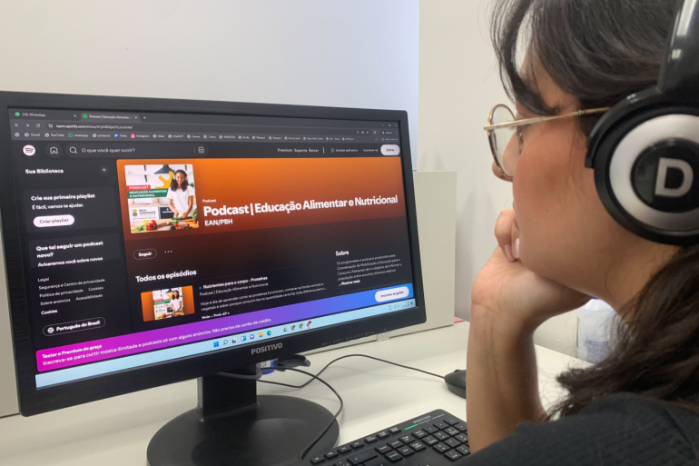 Uma mulher com fones de ouvido de lado para a foto e de frente para um computador que está aberto na página do podcast. 