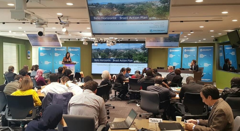 A convite do Banco Mundial, a Secretaria Municipal de Política Urbana foi convidada a representar o Brasil no encontro Technical Deep Dives. Foto: Divulgação PBH Mulher palestra de pé, à esquerda, homem está sentado à direita, quatro mesas com pessoas à frente e vários telões sobre Belo Horizonte, em sala.