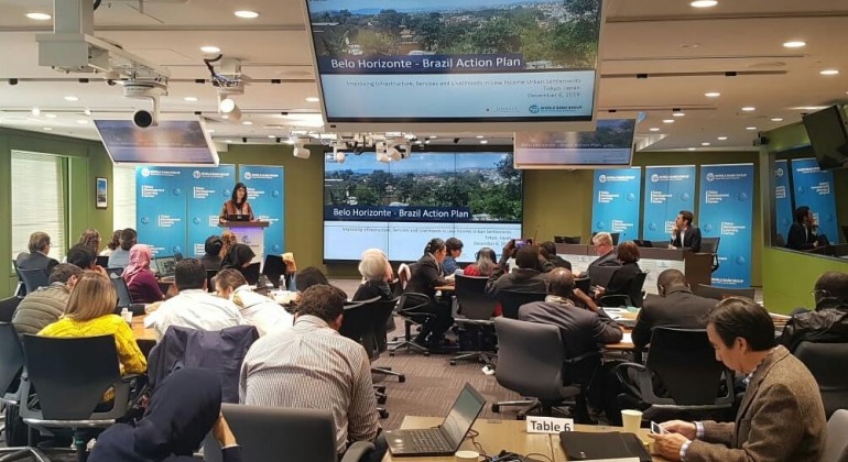 Mulher palestra de pé, à esquerda, homem está sentado à direita, quatro mesas com pessoas à frente e vários telões sobre Belo Horizonte, em sala. 