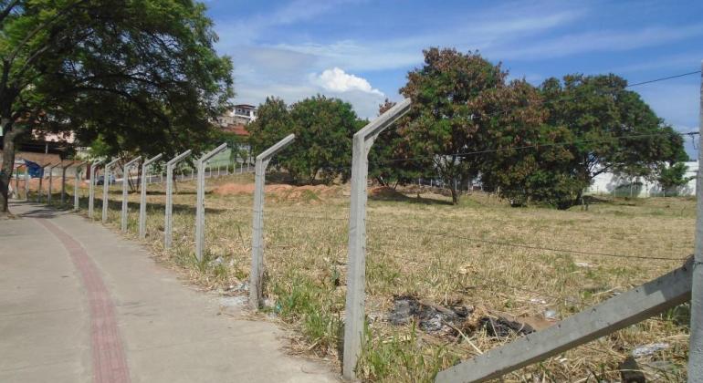 Lote vago com grama aparada e cerca com vigas de cimento e arame farpado ao lado de uma estrada, durante o dia. 