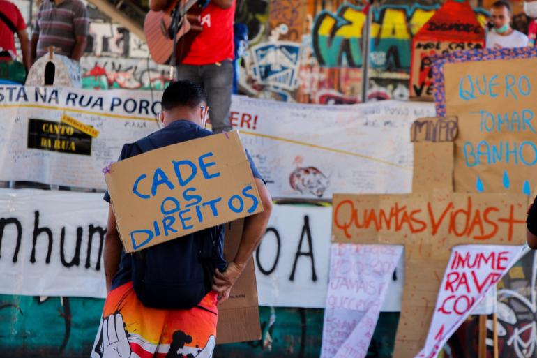 Inscrições abertas até 21 de outubro para eleição suplementar de comitê de políticas para a população em situação de rua A fotografia mostra uma manifestação em defesa dos direitos da população em situação de rua. Em primeiro plano, uma pessoa de costas, com cabelos curtos, camiseta azul-escura, mochila preta e bermuda colorida, carrega nas costas um cartaz de papelão com a frase “Cadê os direitos?”, escrita à mão em letras azuis. Ao fundo, aparecem outras pessoas, faixas e cartazes com reivindicações, incluindo um com a frase “Quero tomar banho”. Uma pessoa segura um violão. O espaço é marcado por paredes cobertas de grafite