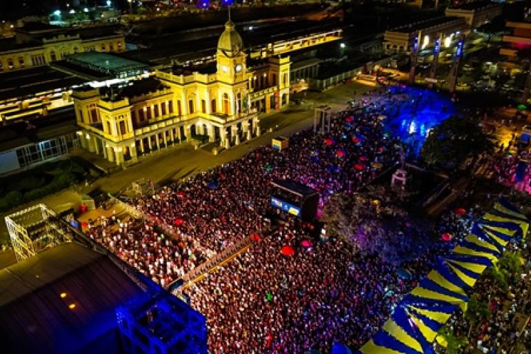 Início da celebração dos 129 anos de BH reúne mais de 20 mil pessoas em shows na Praça da Estação Imagem aérea noturna na Praça da Estação, em Belo Horizonte, tomada por um grande público durante a celebração pelo aniversário de 129 anos da capital mineira. Milhares de pessoas ocupam o espaço em frente ao Museu de Artes e Ofícios, iluminado por tons de amarelo. À direita, estruturas de palco, tendas azuis e amarelas e luzes coloridas compõem o cenário da festa.