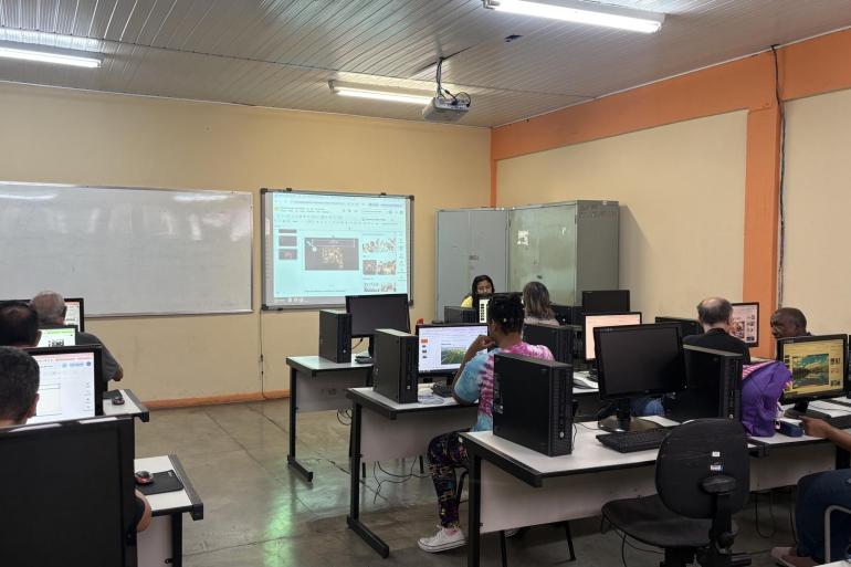 Programa de Inclusão Digital abre inscrições para 200 vagas da oficina gratuita “Descomplicando o gov.br” Laboratório de informática com alunos sentados de costas, cada um focado em um computador de mesa. Na frente da sala, uma instrutora está sentada e há uma tela de projeção e um quadro branco