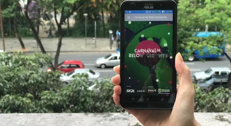 Mão humana segura celular com app Carnava de Belo Horizonte. Ao fundo, árvores e avenida com carros. 