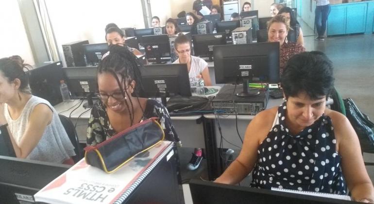 Mais de dez mulheres, sentadas em frente a computadores, com técnico acompanhando, ao fundo. 