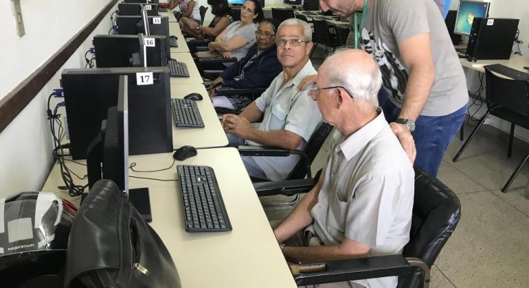 Dois idosos, sentados cada um em frente ao um computador, recebem orientações de monitor, que está de pé. 