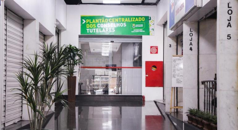 Porte de vidro em galeria comercial, com uma placa verde em cima, com os dizeres: "Plantão Centralizado dos Conselhos Tutelares".