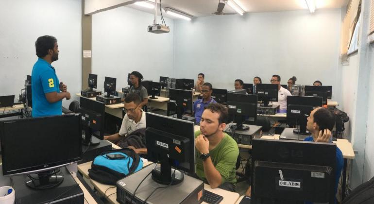 Homem dá aula a cerca de dez alunos, sentados em frente a seus computadores. 