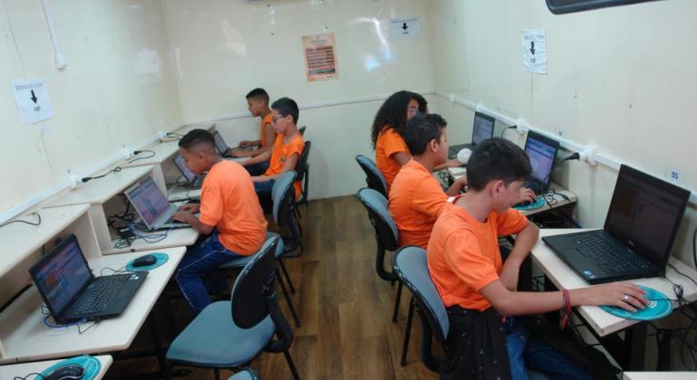 Seis crianças de camiseta laranja usam os computadores da carreta do Projeto Conexão Aberta.