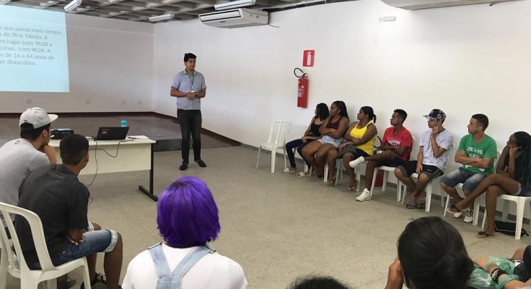 Homem, de pé, dá aula em sala com cerca de treze alunos sentados. 