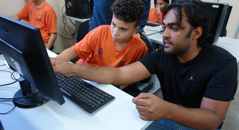 Instrutor jovem ensina aluno de escola municipal. Aluno está sentado e instrutor agachado, ambos em frente a um computador.