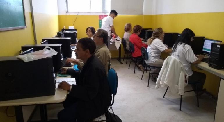 Oito idosos, cada um sentado em frente ao computador, acompanhados por monitor, em sala. 