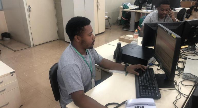 Rapaz sentado em frente ao computador, em uma sala, durante o dia. 