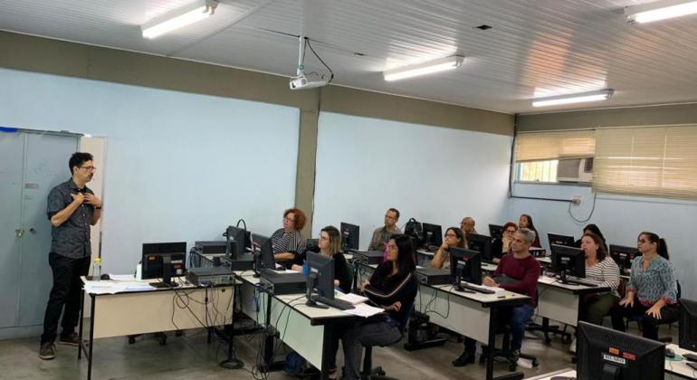 Homem, de pé, palestra para doze pessoas, todas sentadas diante de computadores, em uma sala. 