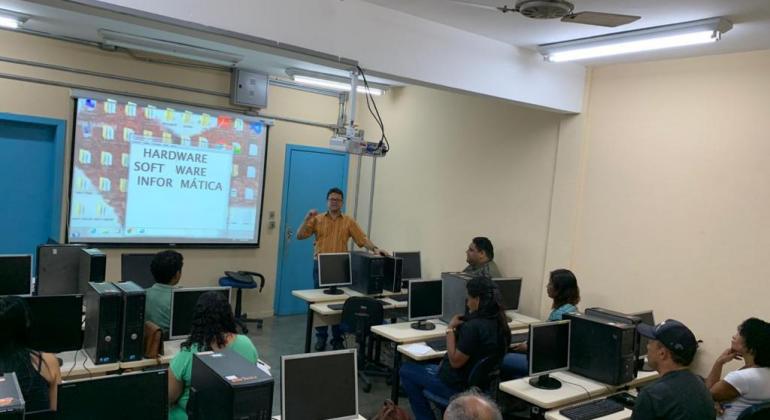 Sete alunos, sentados em suas carteiras, assistem a aula com projetor e professor, em pé, ao lado da projeção. 