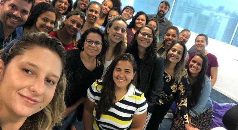 Cerca de vinte mulheres e uns três homens de uma das edições do curso Programando Sonhos Delas posam para foto do tipo selfie, em uma sala. 