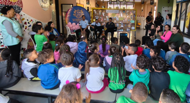 #paratodosverem: na foto, crianças sentadas em fileiras de bancos assistem a uma apresentação musical realizada pela banda da Guarda Municipal. Eles tocam instrumentos e cantam em frente a um mural decorado com desenhos e mensagens. Um banner com o brasão da escola aparece ao fundo, junto a outras pessoas adultas sentadas e em pé, que acompanham e aplaudem a atividade.