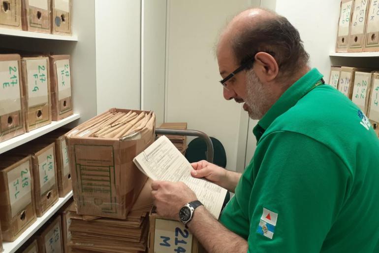 Programa da PBH promove a inclusão no mercado de trabalho de pessoas atendidas na rede de saúde mental Homem de óculos e camisa polo verde examina atentamente um documento em papel impresso dentro de um arquivo de escritório, cercado por várias caixas organizadoras etiquetadas e organizadas em prateleiras.