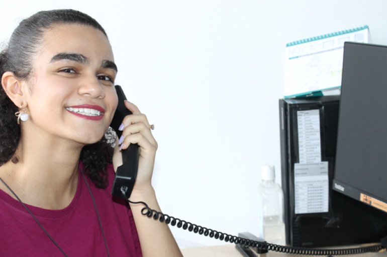 Prefeitura de BH abre 53 vagas de estágio em agosto e bolsas chegam a R$ 1,8 mil Jovem branca com blusa vermelha na frente do computador segurando um telefone