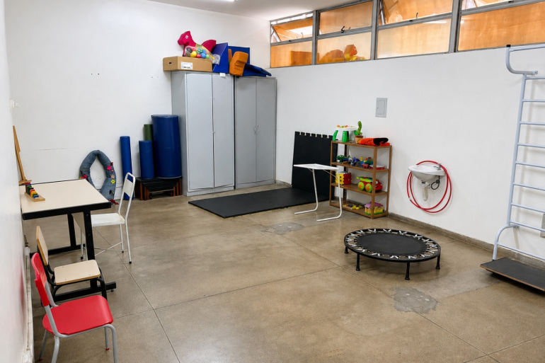 PBH oferece reabilitação especializada para mais autonomia de pessoas com deficiência visual Sala equipada com materiais de reabilitação. Um armário no fundo da sala, uma mesa com cadeiras na lateral esquerda, um trampolim, tatames e brinquedos na lateral direita.