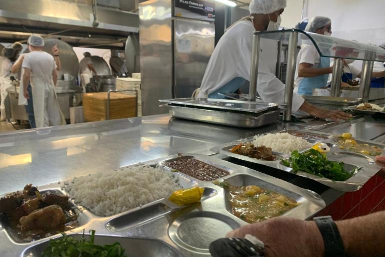 Restaurante Popular de Venda Nova será reaberto ao público nesta segunda-feira (17) após ser reformado Bandeja de metal servida com arroz, feijão, carne e salada em uma linha de atendimento de refeitório industrial, com funcionários de avental e touca ao fundo.