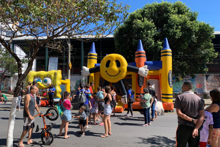 A Rua é Nossa oferece atividades em sete pontos da cidade neste domingo (26) Rua fechada para atividades de lazer, em um dia de céu limpo e ensolarado. Ao centro, brinquedos infláveis recebem crianças, enquanto adultos permanecem ao redor acompanhando a movimentação. Árvores de grande porte fazem sombra sobre parte da via. Ao fundo, há um prédio e um muro com pinturas, compondo um ambiente de convivência, recreação e encontro entre famílias.