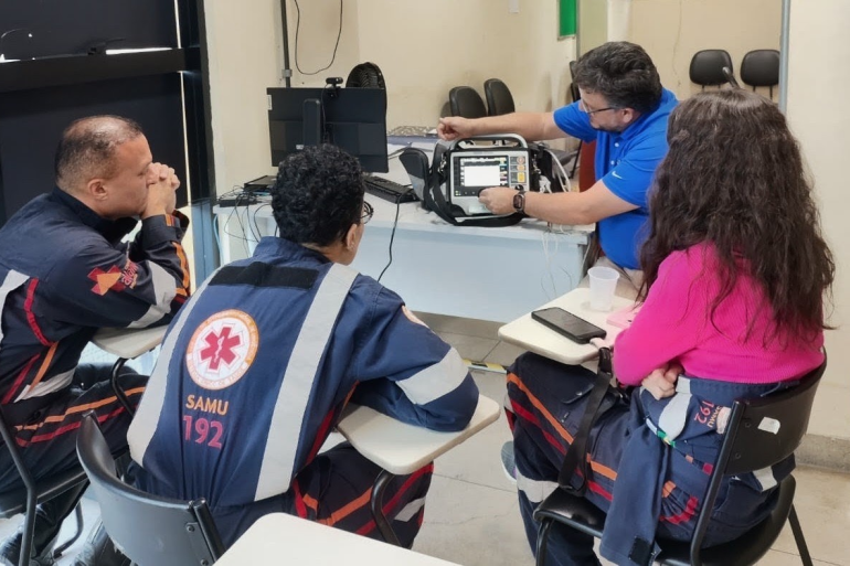 Tecnologia aproxima Samu e hospitais para aprimorar atendimento em casos de infarto Quarto profissionais do Samu, três homens e uma mulher, sentados em carteiras em círculo e um dos homens mostrando um aparelho tecnológico apoiado em cima de uma mesa.