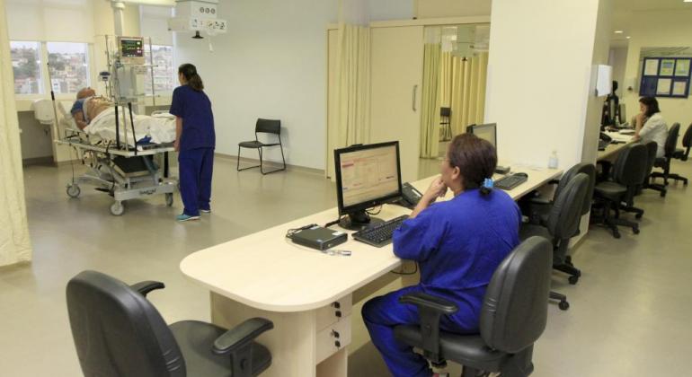 Sala do Hospital Metropolitano Dr. Célio de Castro, com leito hospitalar no qual está deitado um paciente, acompanhado por uma técnica de saúde. À frente, outra técnica consulta o computador. Foto ilustrativa.