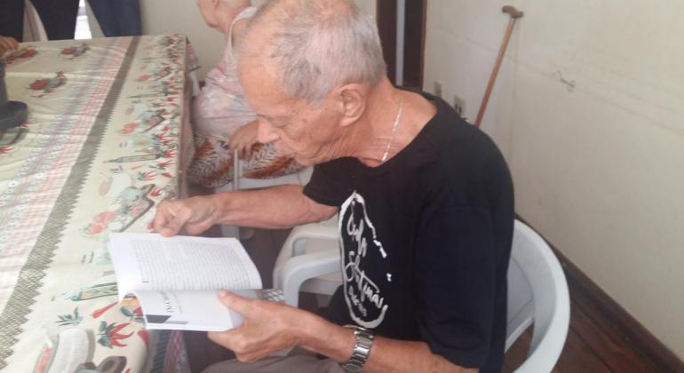 Foto: Karen Moreira/PBH Idoso lê livro, sentado em frente a uma mesa.