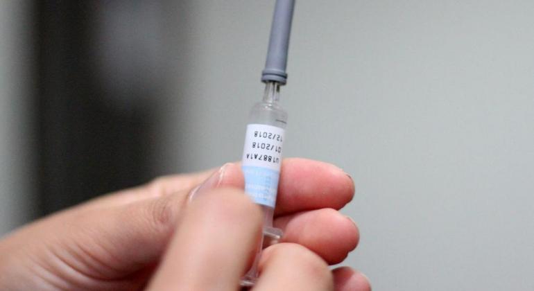 Foto com foco nas mãos de uma pessoa que segura uma ampola de vacina