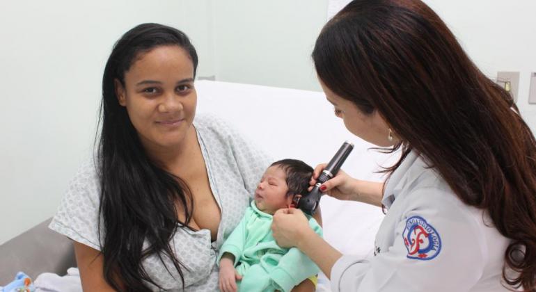 Mãe sefura bebê enquanto técnica de saúde faz teste da orelha. 