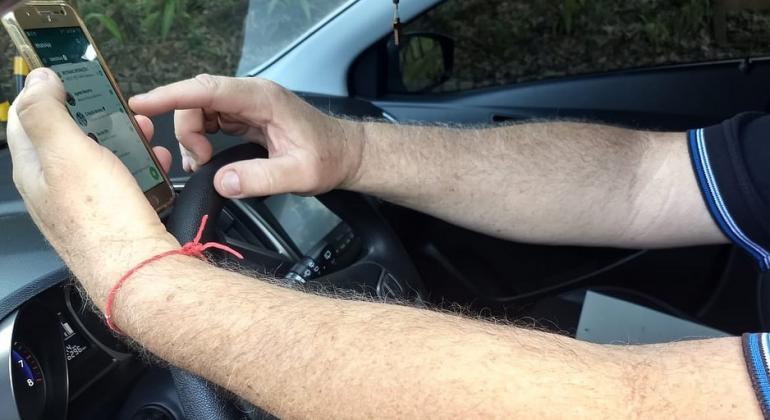 Detalhe de imagem com mãos sobre o volante manipulando um celular.