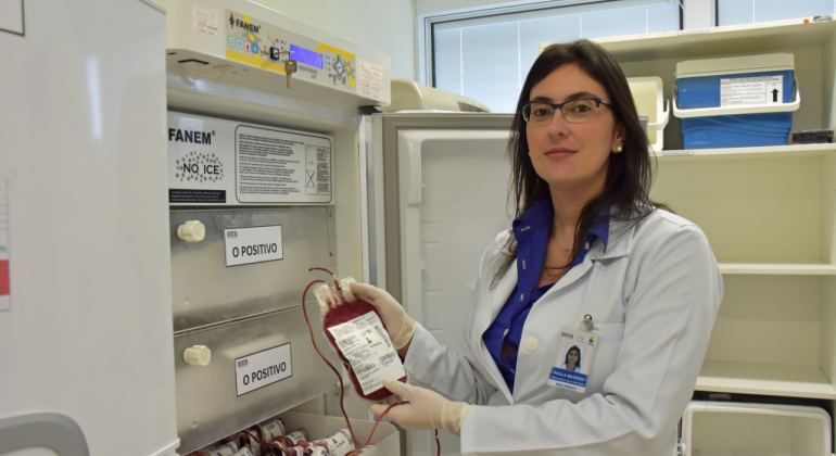 Hospital se tornou, desde 2018, uma das referências na capital mineira para internação de pacientes com complicações da doença falciforme. Foto: Divulgação PBH Mulher com jaleco mostra bolsa de sanque, em frente à geladeira aberta.