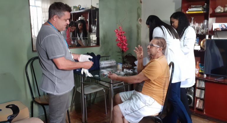 Pacientes que necessitam de cuidados especiais após alta hospitalar têm atendimento domiciliar por meio da PBH. Foto: Leila Porto Homem coloca luvas em frente a outro homem, que está sentado em frente a uma mesa, em uma sala residencial. Ao fundo, duas mulheres vestidas de branco.