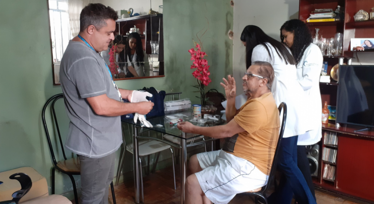 Homem coloca luvas em frente a outro homem, que está sentado em frente a uma mesa, em uma sala residencial. Ao fundo, duas mulheres vestidas de branco. 