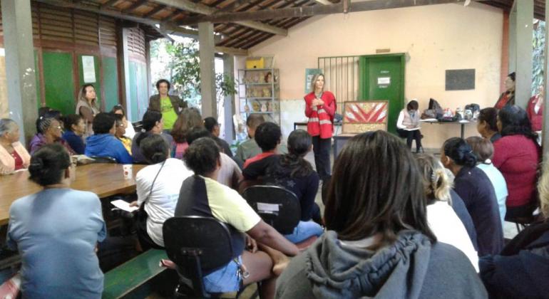 Mulher com colete vermelho dá palestra para cerca de trinta pessoas sentadas. 