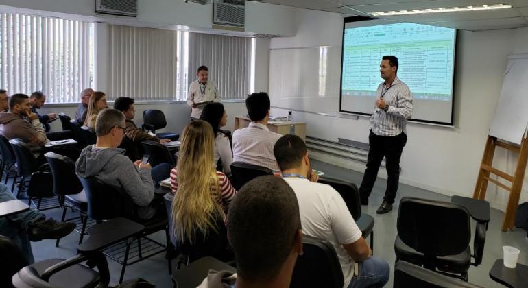 A ação visa qualificar agentes públicos de saúde para que adotem comportamentos seguros durante o manuseio de materiais perfurocortantes. Foto: Divulgação PBH Homem dá palestra para cerca de dez pessoas sentadas, com telão ao fundo.