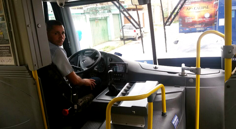 Imagem interna de ônibus, com motorista parado, olhando para trás. 