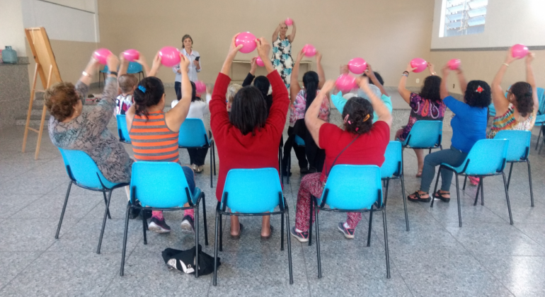Palestra com o tema “Violação de Direitos da Pessoa Idosa” foi tema de roda de conversa no Centro de Saúde São José em junho. Foto: Divulgação PBH Mais de dez pessoas, sentadas, cada uma erguendo uma bola rosa entre as mãos, e uma mulher atrás, fazendo o mesmo movimento.