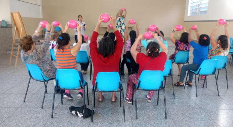 Mais de dez pessoas, sentadas, cada uma erguendo uma bola rosa entre as mãos, e uma mulher atrás, fazendo o mesmo movimento.